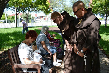 Two Friars standign outside bending down to two seniors seated on a bench